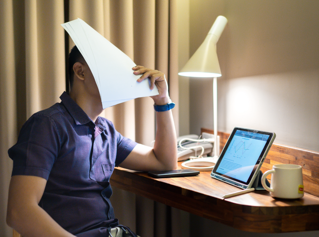 Photo of a man covering his face with papers in frustration or boredom. He is sat a desk with an open laptop, on the screen is a non-descript line graph.