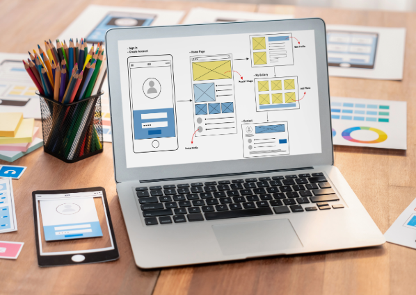Picture of a screen with a mock website. Papers on the desk illustrate website development criteria such as mobile first layout, colour palettes, layouts